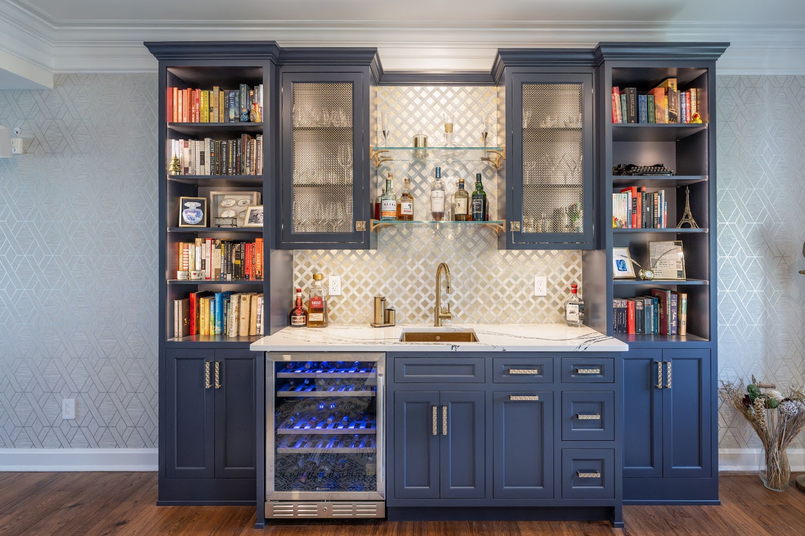 A home wet bar by Laslo Custom Kitchens. Glass shelves in the middle with glasses and bottles of alcohol. Cabinets to the right and left are navy with metal mesh inlay. Navy bookshelves to the right and left. Stainless steel cooler below sink and to the left of navy cabinets and drawers. Backsplash is metallic squares.