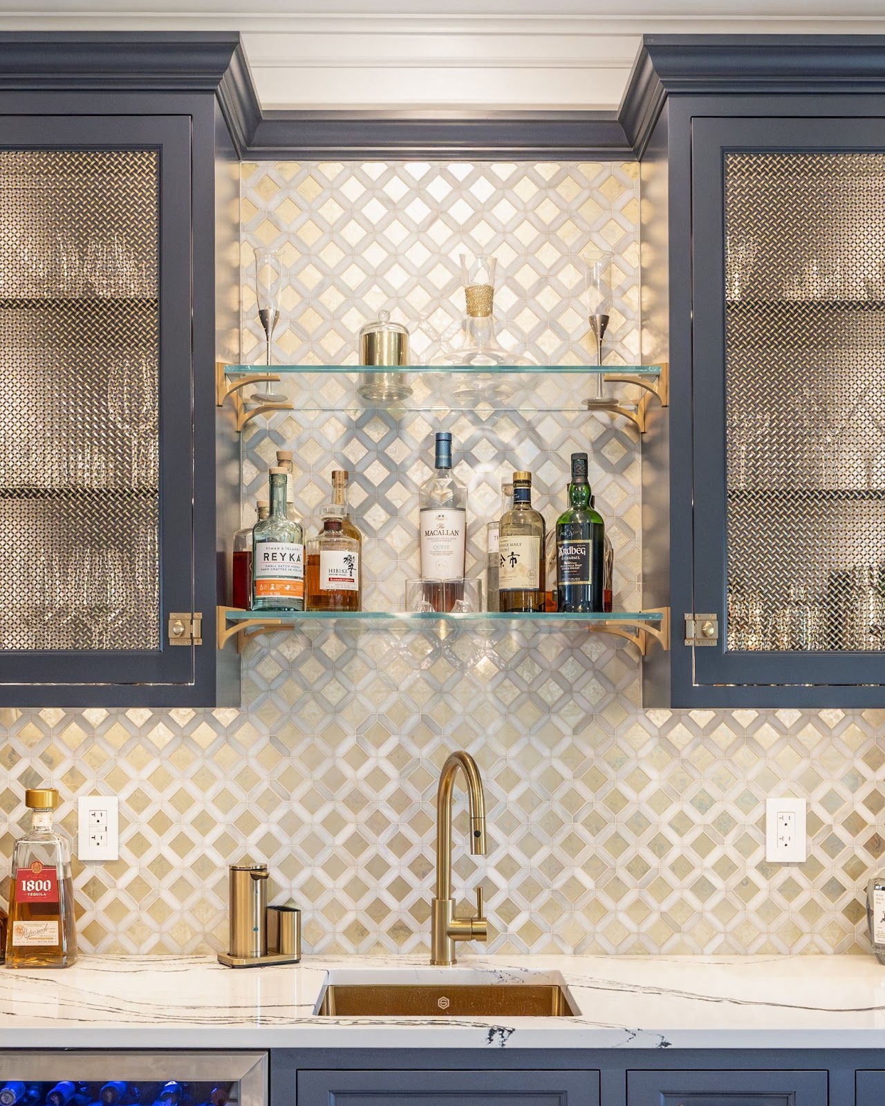 Closeup of a home wet bar by Laslo Custom Kitchens. Glass shelves in the middle with glasses and bottles of alcohol. Cabinets to the right and left are navy with metal mesh inlay. Backsplash is metallic squares.
