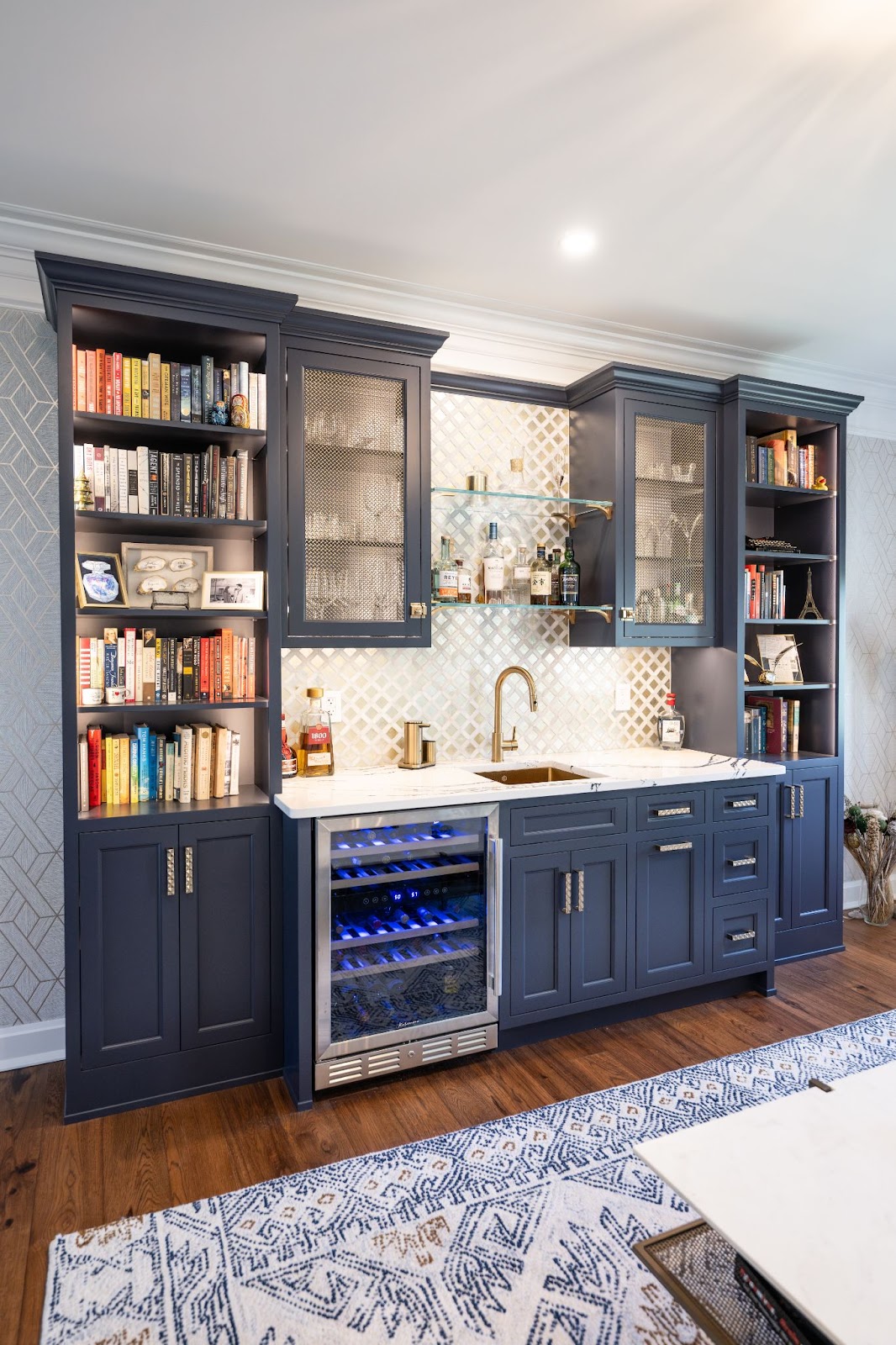 A home wet bar by Laslo Custom Kitchens. Glass shelves in the middle with glasses and bottles of alcohol. Cabinets to the right and left are navy with metal mesh inlay. Navy bookshelves to the right and left. Stainless steel cooler below sink and to the left of navy cabinets and drawers. Backsplash is metallic squares.