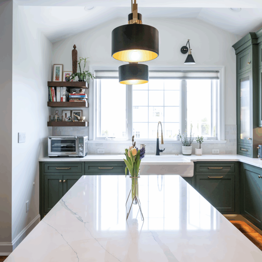 A kitchen with green cabinets by Laslo Custom Kitchens and black pendant lights over a large island with white counter.