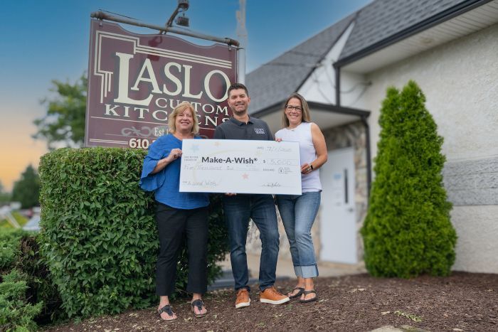 Two women and a man stand in front of a brown Laslo Custom Kitchens sign with a large check for Make-A-Wish.