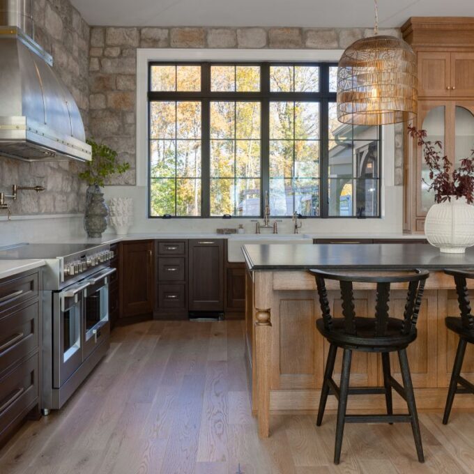 A gorgeous rustic-modern kitchen showcasing the possibilities of funding with a smart kitchen loan, featuring a natural stone accent wall, a light wood island topped with luxurious Cambria Quartz, and a large window overlooking autumn trees.