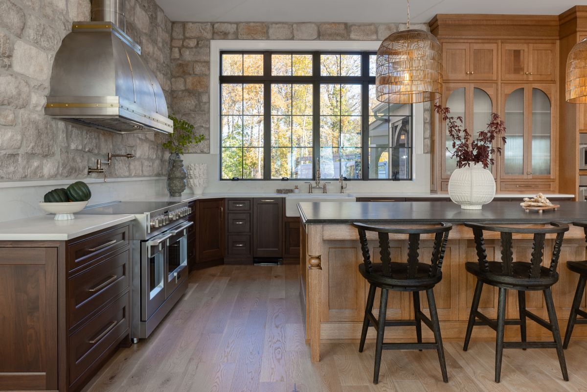 A gorgeous rustic-modern kitchen showcasing the possibilities of funding with a smart kitchen loan, featuring a natural stone accent wall, a light wood island topped with luxurious Cambria Quartz, and a large window overlooking autumn trees.