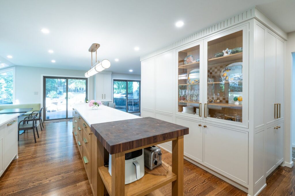 Glass-front taupe kitchen cabinet with wood shelves and fluted crown beside the island’s wood-topped extension.