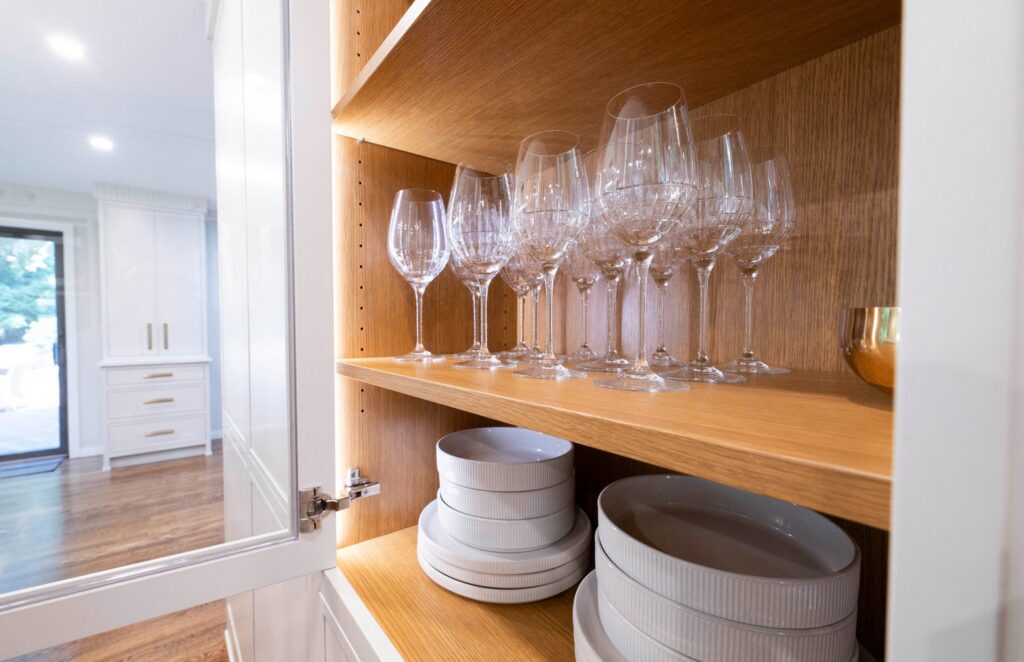 Open glass-front kitchen cabinet with a wood interior, wine glasses, and stacked dishes.